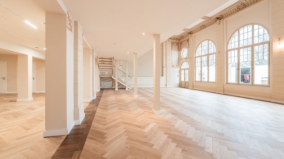 Großer, leerer Innenraum mit hellem Fischgrätparkett, vielen weißen Säulen und offener Treppe im Hintergrund. An der rechten Wand befinden sich hohe Rundbogenfenster und dekorativer Stuck unter einer hohen Decke.