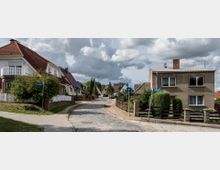 Straßenszene in einer Wohnsiedlung mit Einfamilienhäusern, teils mit roten Ziegeldächern und weißen Fassaden sowie gepflegten Gärten. Die Straße ist mit Kopfsteinpflaster versehen, flankiert von Bürgersteigen und Straßenschildern, unter einem bewölkten Himmel.