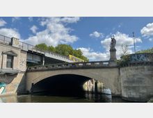 Brücke mit einer Steinfigur auf einem Pfeiler, darunter ein Wasserkanal. Auf der oberen Brücke fährt eine gelbe U-Bahn, daneben ist ein kleines Gebäude mit beschmierter Fassade zu sehen.