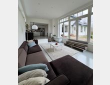 Helles, modernes Wohnzimmer mit großem braunem Ecksofa, weißem Teppich und kleinem Couchtisch, das offen in eine Küche übergeht. Bodentiefe Fensterfront mit seitlichen Stufen führt zur Terrasse und bietet Blick in den Garten und auf ein angrenzendes Haus.