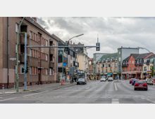 Städtische Straßenszene mit mehrspuriger Fahrbahn und umliegenden Gebäuden, darunter Wohn- und Geschäftshäuser. Rechts und links der Straße befinden sich Ladenzeilen, während mehrere Autos und Fußgänger im Bereich der Kreuzung zu sehen sind; Ampelanlagen und Straßenschilder sind vorhanden.