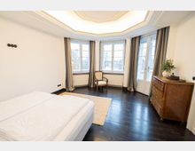 Helles Schlafzimmer mit dunklem Holzboden, abgehängter Decke mit indirekter Beleuchtung und einer Fensterfront aus mehreren großen, bodennahen Fenstern mit Vorhängen. Rechts steht eine Holzkommode neben einer verglasten Tür zum Balkon.