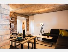 Ein modernes Wohnzimmer mit offener Backsteinwand und Gewölbedecke, eingerichtet mit einem Esstisch, gedecktem Geschirr und Gläsern. Im Hintergrund stehen eine schwarze Sofalandschaft, ein Couchtisch, eine große Stehlampe und Zimmerpflanzen.