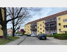 Wohnstraße mit einem mehrstöckigen Wohnhaus in Gelb und Lila. Vor dem Haus stehen geparkte Autos, ein Gehweg verläuft entlang der Straße, und im Vordergrund sind kahle Bäume sowie kleinere Grünflächen zu sehen.