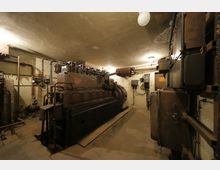 Ein technischer Raum mit einem großen, alten Generator oder Dieselmotor in der Mitte. Sichtbar sind Rohre, Schaltkästen und Metallstrukturen an den Wänden, mit spärlicher Beleuchtung und einer unverputzten Decke.