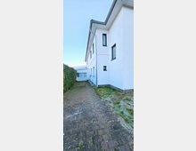 Einfamilienhaus mit weißer Fassade und Flachdach, daneben ein gepflasterter Weg, der entlang einer Hecke führt. Im Vordergrund sind gerahmte Fenster und ein leicht zugefrorener Rasen erkennbar, während im Hintergrund ein Nebengebäude sichtbar ist.