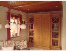 Wohnzimmer mit Holzmöbeln, einschließlich eines großen Schranks mit Glas- und Holztüren. Im Vordergrund steht ein gepolstertes Sofa mit floralem Muster, darüber hängt eine Deckenlampe mit mehreren kugelförmigen Leuchten. Ein Fenster mit roten Vorhängen und Pflanzen auf der Fensterbank befindet sich links im Raum.