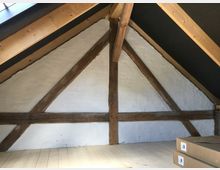 Ansicht eines Dachbodens mit Holzgebälk und weiß verputzten Wänden. Der Raum hat ein steiles Satteldach, sichtbare Holzbalken und einen hellen Holzfußboden; im Vordergrund liegen zwei Kartons.