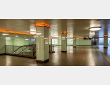 U-Bahn-Station mit verzierter, moderner Innenarchitektur. Glänzende Säulen, orange Beleuchtung an den Deckenrändern, mehrere Rolltreppen und Treppen führen zu den Bahnsteigen.
