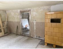Ein leerer, renovierungsbedürftiger Raum mit einer beschädigten Wand, einer offenen Tür zu einem weiteren unordentlichen Bereich und einem gelblich-braunen, gefliesten Kachelofen auf der rechten Seite. Der Boden besteht aus einfachem Beton, und einzelne Holzrahmenteile sind an der Wand angelehnt.