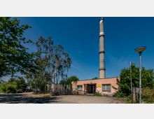 Industrieanlage mit einem hohen Schornstein im Zentrum, einem flachen rosa Gebäude im Vordergrund und umgeben von Bäumen. Im Außenbereich sind Fahrräder, parkende Autos und ein Metallzaun sichtbar.