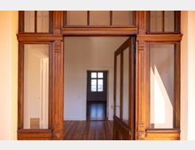 Holzrahmen einer offenen Tür mit dekorativen Verzierungen, die in einen Innenraum mit Holzboden und weißen Wänden führt; durch einen weiteren Türrahmen ist ein kleiner Raum mit einem Fenster sichtbar.