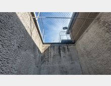 Blick nach oben auf einen Außenbereich mit hohen Betonwänden, bedeckt von einem Maschendrahtzaun, durch den der Himmel sichtbar ist. Im oberen Bereich befinden sich Geländer und ein kleines Podest, das Teil einer Metallkonstruktion ist.