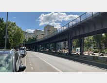 Eine erhöhte Stahlbahntrasse verläuft parallel zu einer Straße mit parkenden Autos. Im Hintergrund befinden sich Bäume, moderne Gebäude und eine Baustelle mit Absperrzäunen.