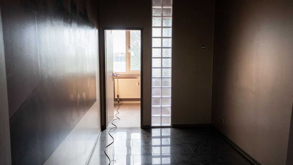 Schmaler, leerer Innenflur mit glänzendem Fliesenboden und glatten Wänden, am Ende eine offene Tür zu einem hellen Raum mit Fenster und Heizkörper. Rechts neben der Tür verläuft ein vertikales Band aus Glasbausteinen, und ein Kabel liegt über den Boden in Richtung Tür.