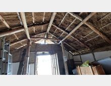 Innenraum eines traditionellen Gebäudes mit einem Dach aus Holzträgern und Stroh. Die Wände bestehen aus Lehm und Holz, und durch eine geöffnete Holztür fällt Tageslicht in den Raum, in dem sich auch Leitern und Holzkisten befinden.