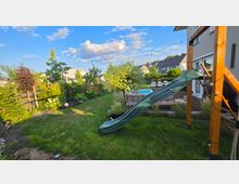 Blick in einen gepflegten Wohnhausgarten mit Rasenfläche, Beeten und mehreren Bäumen; rechts steht eine Holzterrasse am Haus mit Sitzgruppe und kleinem Pool. Im Vordergrund führt eine grüne Rutsche von einer Holzspielkonstruktion in den Garten, darüber sind Lichterketten gespannt.