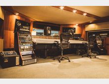 Ein Tonstudio mit einer großen Mischkonsole in der Mitte, umgeben von Lautsprechern, Reglern und Racks mit Audioequipment. Der Raum hat hölzerne Wandverkleidungen, einen Teppichboden und gedämpfte Beleuchtung.