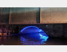 Ein Tunnel mit gemauertem Bogen, dessen Umrandung mit blauem Licht beleuchtet ist. Der Boden des Tunnels ist mit Wasser bedeckt, in dem sich das Licht spiegelt.