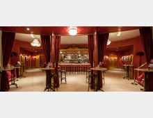 Ein eleganter Barbereich mit roter Wand- und Deckenverkleidung, goldenen Akzenten und Kronleuchtern. Im Vordergrund stehen hohe Tische mit Barhockern, im Hintergrund eine Bar mit Glasregalen voller Flaschen, eingerahmt von schweren roten Vorhängen.