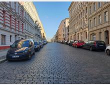 Straße mit Kopfsteinpflaster, gesäumt von parkenden Autos und mehrstöckigen Altbauten mit verzierten Fassaden. Der klare Himmel und das Licht betonen die detaillierte Architektur der Gebäude.