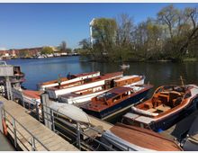 Boote im Hafen an einem Flussufer, mit mehreren weißen und hölzernen Motorbooten, die an einem Steg festgemacht sind. Im Hintergrund sind Bäume, ein Industriebau mit einem Schornstein sowie ein ruhiges Gewässer mit einem kleinen Ruderboot zu sehen.