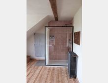 Badezimmer mit einer Eckdusche, die durch eine Glaswand mit schwarzem Rahmen abgetrennt ist, rosa Fliesen an der Duschwand, einem hölzernen Boden und einer sichtbaren Holz-Deckenbalken. Ein Handtuchhalter mit Handtüchern, ein schwarzer Heizkörper und ein geflochtener Wäschekorb sind ebenfalls im Raum.