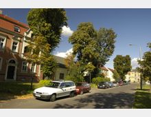 Eine ruhige Wohnstraße mit geparkten Autos am Straßenrand. Im Hintergrund sind mehrstöckige Wohnhäuser mit roten Ziegeldächern sowie grüne Bäume zu sehen, unter einem klaren blauen Himmel.
