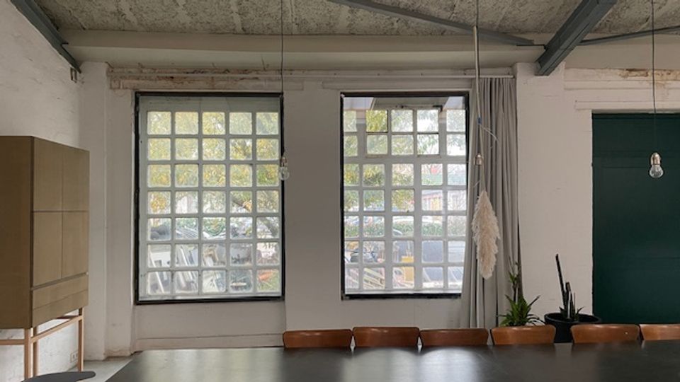 Industrieartiger Innenraum mit hoher Betondecke und sichtbaren Trägern, zwei großen Rasterfenstern und einem langen Tisch mit Stühlen im Vordergrund. Rechts befindet sich eine dunkle Tür sowie hängende Glühbirnen und einige Pflanzen.