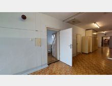 Ein einfacher Innenflur mit beigen Wänden und braun-orangem Bodenbelag. Eine geöffnete Tür führt in einen Raum mit einem gepolsterten Türrahmen, im Flur stehen Schränke und Türen, und an der Decke befinden sich Leuchtstofflampen.