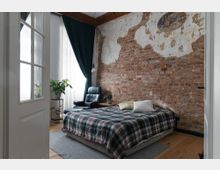 Schlafzimmer mit einer freiliegenden Backsteinwand, einem Bett mit kariertem Überwurf, einem schwarzen Ledersessel und Vorhängen vor großen Fenstern. Der Raum ist mit Holzboden, einem kleinen Beistelltisch und Pflanzen dekoriert.