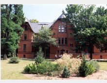 Rotes Backsteingebäude im neugotischen Stil mit spitzem Giebeldach und hohen, bogenförmigen Fenstern, umgeben von Bäumen und einem gepflegten Garten mit Sträuchern und Grasflächen. Im Vordergrund ist eine Gruppe von Menschen nahe dem Eingang zu sehen.