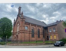 Backstein-Kirche mit neogotischer Architektur, darunter Spitzbogenfenster und Rundfenster, flankiert von Bäumen und umgeben von einem schmiedeeisernen Zaun. Im Vordergrund verläuft ein Bürgersteig, und ein geparktes Auto steht an der Straße.