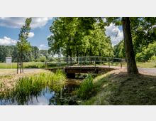 Eine kleine Brücke mit Metallgeländer überspannt einen schmalen Bach in einer parkähnlichen Umgebung mit grünen Wiesen und Bäumen. Im Hintergrund sind ein Weg, ein Schild und dichter Wald erkennbar, während der Himmel von weißen Wolken bedeckt ist.