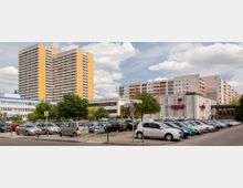 Parkplatz mit geparkten Autos im Vordergrund, im Hintergrund mehrere Hochhäuser mit orangefarbenen und beigen Fassaden. Rechts ein Gebäude mit der Aufschrift "Kino Sojus", umgeben von Bäumen und kleineren Gewerbebauten.