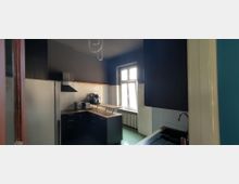 Schmale Küche mit dunklen Wänden, weißen Fliesen und schwarzen Unterschränken mit Holzarbeitsplatte. Ein großes Fenster über einem Heizkörper sorgt für Tageslicht; sichtbar sind zudem Kühlschrank, Dunstabzugshaube und Spüle.