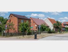 Ruhige Wohnstraße mit freistehenden Einfamilienhäusern aus rotem Backstein und Satteldächern, umgeben von kleinen Vorgärten mit Bäumen und niedrigem Holzzaun. Im Vordergrund befinden sich eine Straßenlaterne, ein Abfallbehälter und Verkehrsschilder.