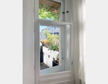 Blick durch ein geöffnetes, weißes Fenster auf einen kleinen Balkon mit Pflanzen in Töpfen und Kletterpflanzen an einem Holzgitter; im Hintergrund sind moderne Wohngebäude sichtbar.