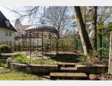 Gartenbereich mit einem runden Pavillon aus Metall und einem Tisch mit Stühlen darunter, umgeben von niedrigen Büschen und großen Bäumen. Im Hintergrund befinden sich Wohnhäuser mit roten Ziegeldächern.