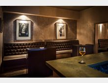 Eine stilvolle Lounge mit braunen, gepolsterten Lederbänken entlang der Wand, beleuchtet durch warme, indirekte Beleuchtung. An der Wand hängen zwei schwarz-weiße Fotografien, und ein Holztresen im Vordergrund trägt ein Glas mit einem Getränk.