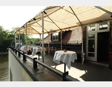 Überdachte Dachterrasse eines Restaurants mit weißen Tischdecken auf runden Tischen und schwarzem Geländer entlang des Randes. Rechts sind Dachziegel, Fenster und eine Glastür des Gebäudes zu sehen, links blickt man auf Bäume und ein Gewässer darunter.