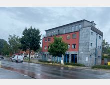 Mehrstöckiges Gebäude an einer nassen Straße bei bewölktem Himmel, mit einer Fassade aus grauem Stein und roten Wandelementen. Vor dem Gebäude stehen mehrere Bäume, und im Hintergrund sind kleinere Gebäude sowie Verkehrsschilder zu sehen.