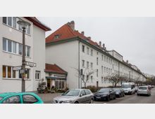 Straßenansicht einer Wohnsiedlung mit mehrstöckigen, weißen Gebäuden und roten Ziegeldächern. Am Gehweg sind ein Blumenladen, parkende Autos und Straßenschilder zu sehen, im Hintergrund stehen kahle Bäume unter einem grauen Himmel.