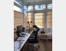 Ein heller Arbeitsbereich in einem verglasten Raum mit mehreren Fenstern, die von beigefarbenen Rollos bedeckt sind. Links steht ein Schreibtisch mit Monitor, Laptop und Büromaterial, rechts ein Sofa mit dekorativen Kissen, daneben ein kleiner Hocker.