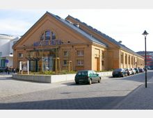 Backsteinbau mit der Aufschrift „Hans Otto Theater“ über dem Eingang, ergänzt durch große Fenster und ein vorgelagertes Glasvordach. Vor dem Gebäude ist ein kleiner Parkplatz sowie eine gepflasterte Straße zu sehen.