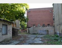 Verlassener Hinterhof mit heruntergekommenen Gebäuden, einer Backsteinmauer und von Graffiti bedeckten Wänden. Im Vordergrund ist ein betonierter Weg, umgeben von überwuchertem Grün und Ästen, während im Hintergrund ein großes rotes Ziegelgebäude zu sehen ist.