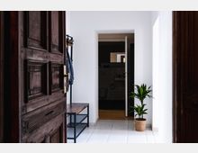 Blick in einen hellen Flur mit weißen Wänden und Fliesenboden. Links steht eine geöffnete, dunkle Holztür, daneben ein Garderobenständer mit Kleidungsstücken und eine Sitzbank, rechts steht eine grüne Zimmerpflanze in einem Korb; im Hintergrund ist ein Badezimmer sichtbar.