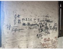 Eine helle Ziegelwand mit handschriftlichen Inschriften in dunkler Tinte, darunter Namen und Daten wie „19.3.25“. Im unteren Bereich ist unverputztes Mauerwerk sichtbar, und rechts befindet sich ein Fensterrahmen mit Holz und Glas.