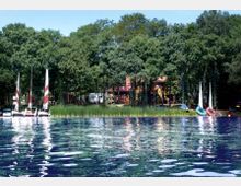 Seeufer mit mehreren Segelbooten auf dem Wasser, im Hintergrund dichter Wald und ein Gebäude, das teilweise zwischen den Bäumen sichtbar ist. Links und rechts der Boote befinden sich weitere kleine Wasserfahrzeuge.