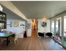 Wohnbereich in einem modernen Innenraum mit heller Holzfußboden, einer langen Tafel mit unterschiedlichen Stühlen und einer Galerie aus Dekorplatten an der Wand. Auf der rechten Seite befinden sich große Glasfenster, die zu einem Balkon führen.